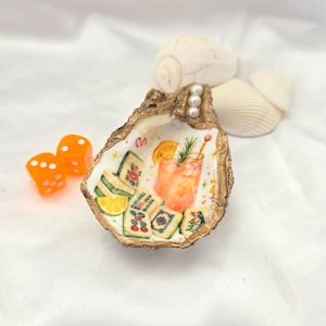 May include: A decorative oyster shell dish with a gold-painted rim and three small white pearls. The interior features a painted illustration of a cocktail, mahjong tiles, and a lemon slice. Two bright orange dice are positioned to the left.