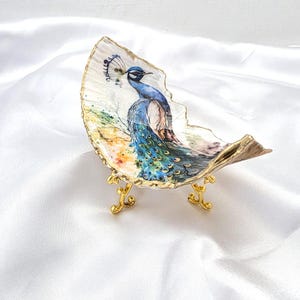 May include: A decorative oyster shell dish featuring a colorful peacock illustration with blue, green, and gold tones. The shell has a gilded gold edge and is supported by a small, ornate gold-colored metal stand with three curved legs.