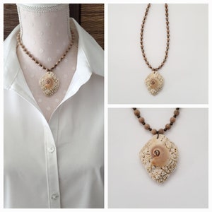 May include: A brown wooden bead necklace with a seashell pendant. The seashell is a light brown color and has a spiral pattern. The necklace is displayed on a white mannequin.