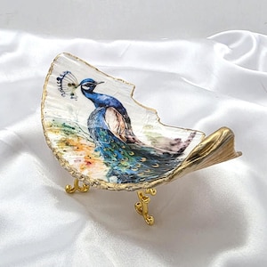 May include: A curved, shell-shaped decorative dish featuring a detailed illustration of a peacock with vibrant blue and green plumage. The edges of the dish are finished with a metallic gold trim, and it stands on three ornate, gold-colored metal legs.