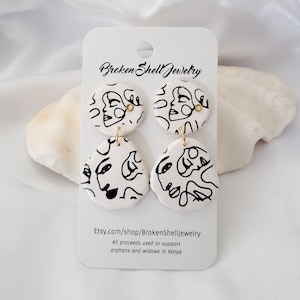 May include: White ceramic earrings with black line drawings of faces. The earrings are a round shape and a freeform shape. The earrings are on a white card with the text "Broken Shell Jewelry" and "Etsy.com/shop/BrokenShellJewelry".