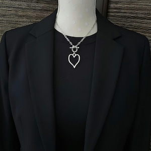 May include: A silver-toned necklace featuring a chain with a toggle clasp and a heart-shaped pendant. The necklace is displayed against a black shirt and a black blazer. The heart pendant has an open center design and hangs from the toggle closure.