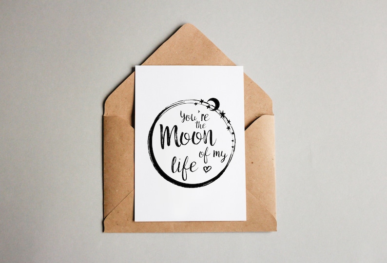 Puede incluir: Una tarjeta blanca con un c&iacute;rculo negro y el texto "You're the Moon of my life" dentro del c&iacute;rculo. La tarjeta est&aacute; en un sobre marr&oacute;n.