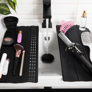 May include: Black sink tidies holding beauty products and hair tools. Includes makeup brushes, curling tongs, hairbrush, and cosmetics. A black collapsible cup and a plant are also visible.
