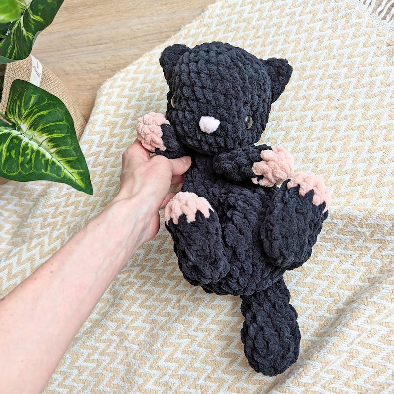 May include: A black crocheted cat plush toy with pink paw pads. The toy is lying on a white and beige patterned fabric.