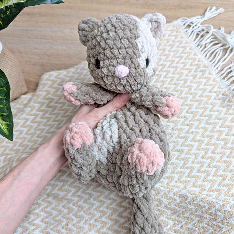 May include: A crocheted plush toy cat in shades of gray and pink. The cat has a long tail and pink accents on its paws and nose.