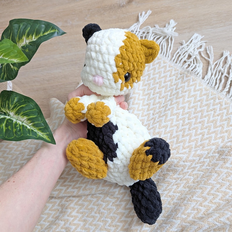 May include: A crocheted cat toy with white, black, and orange yarn. The cat is lying on a white and beige blanket.