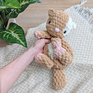 May include: A crocheted cat toy with brown and white fur and pink accents. The cat is lying on its back with its paws in the air.