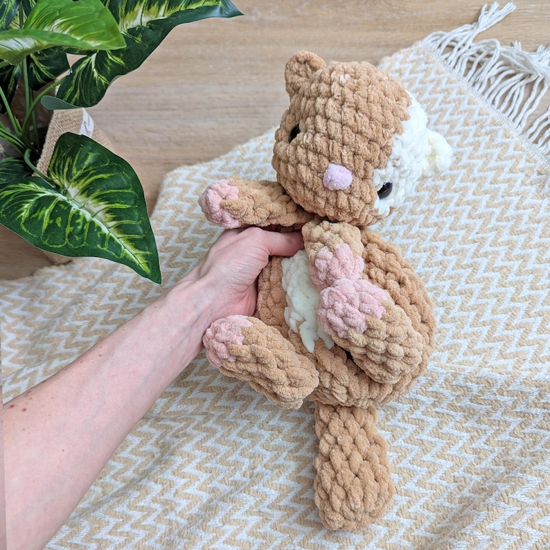 May include: A crocheted cat toy with brown and white fur and pink accents. The cat is lying on its back with its paws in the air.