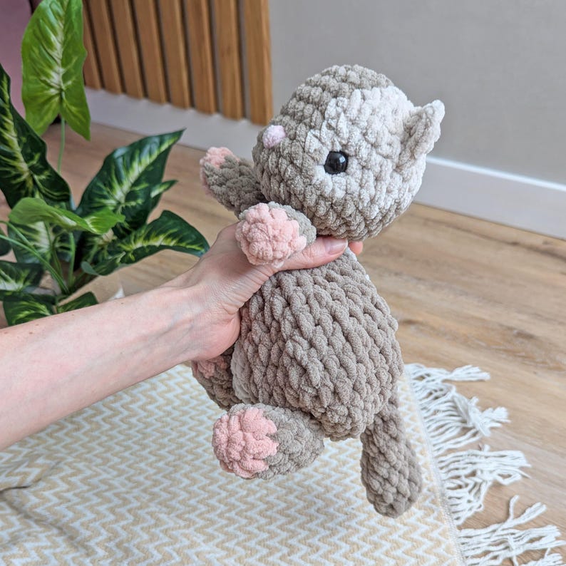 May include: A crocheted plush toy cat in brown and white yarn with pink accents. The cat has a white face with black eyes and a pink nose.
