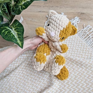 May include: A crocheted plush toy cat with a cream and mustard yellow color scheme. The cat is lying on a beige and white chevron patterned blanket.