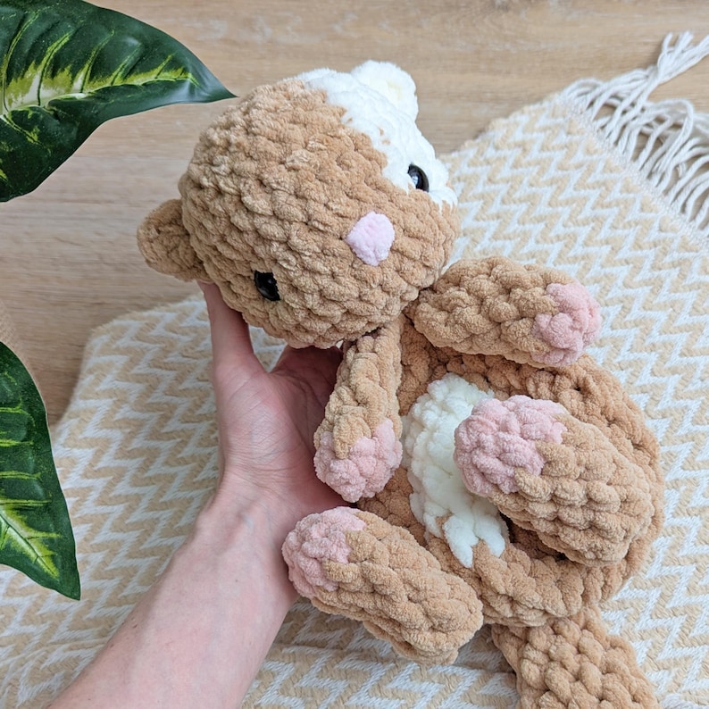 May include: A crocheted stuffed animal cat with brown and white fur and pink accents. The cat is lying on its side with its paws visible.