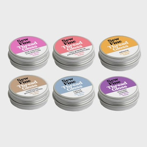 Könnte beinhalten: Sechs runde silberne Metalldosen mit Sew Fine Thread Gloss, die jeweils 15 ml oder 0,5 oz Produkt enthalten. Die Etiketten sind in verschiedenen Farben wie Pink, Rot, Orange, Beige, Blau und Lila gehalten und tragen Namen wie Pink Macaron und Lavendel.