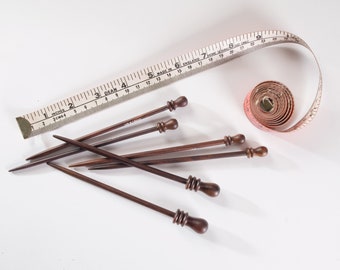 May include: A set of six wooden knitting needles with brown and black tips. The needles are arranged in a starburst pattern on a white surface. A white tape measure with black markings is also visible in the image.