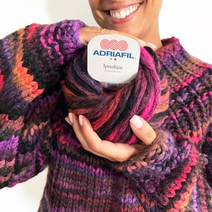 May include: A person wearing a multi-colored striped sweater holding a skein of yarn labeled "ADRIAFIL Ipnotico".
