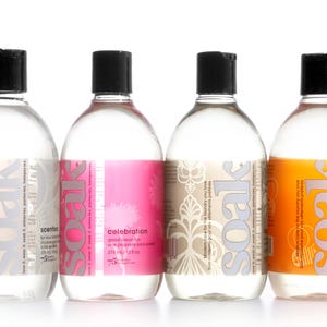 Op de afbeelding: Vier doorzichtige plastic flessen met wasmiddel met witte etiketten en zwarte doppen. De etiketten hebben het woord "soak" erop gedrukt in een gestileerd lettertype. De flessen zijn in een rij van links naar rechts gerangschikt. De flessen zijn gelabeld met "geparfumeerd", "viering", "origineel" en "citrus".