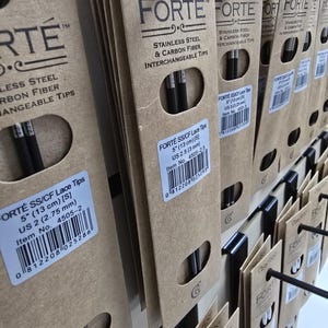 Könnte beinhalten: Nahaufnahme von mehreren Packungen austauschbarer Stricknadelspitzen. Die Verpackungen bestehen aus braunem Karton und tragen den Markennamen "FORTE" sowie Produktinformationen. Die Spitzen sind schwarz und bestehen aus Edelstahl und Kohlefaser.