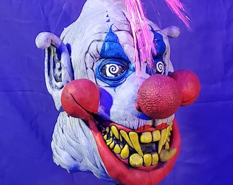 May include: A creepy clown mask with a large, toothy grin, red nose, and blue and white makeup. The mask has a pink and white fiber optic hairpiece and is mounted on a black stand.