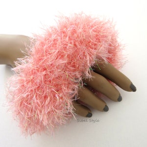 Puede incluir: Un brazalete de lana esponjosa en color rosa coral, elaborado con hilo texturizado y fibras iridiscentes entrelazadas. El accesorio presenta una apariencia densa y peluda con un acabado suave que refleja la luz. El texto Sibel Style est&aacute; impreso en negro.