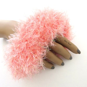 Puede incluir: Un pu&ntilde;o de mu&ntilde;eca de color rosa claro, difuso y hecho de hilo texturizado con fibras iridiscentes tejidas. El accesorio presenta una apariencia densa y peluda, exhibido en una mano de maniqu&iacute; con el texto Sibel Style en el dedo &iacute;ndice.