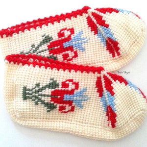 May include: A pair of cream-colored crochet slippers featuring a red, green, and light blue floral pattern. The slippers have a red crocheted trim along the top edge and the text Sibel Style is printed in small black letters on the side.