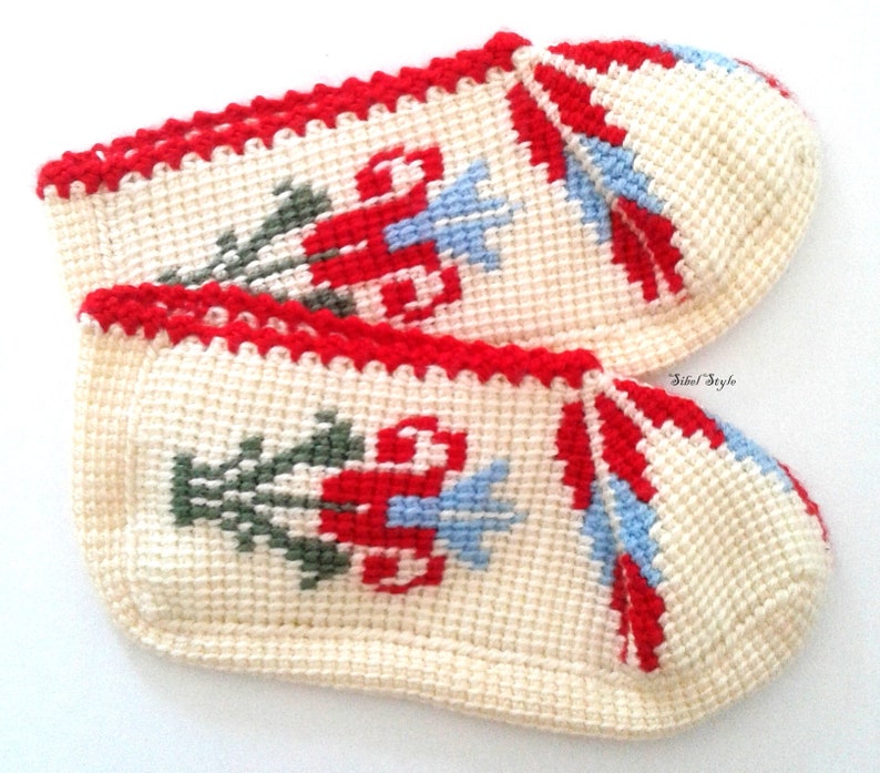 May include: A pair of cream-colored crochet slippers featuring a red, green, and light blue floral pattern. The slippers have a red crocheted trim along the top edge and the text Sibel Style is printed in small black letters on the side.