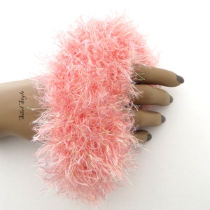 Puede incluir: Un coletero circular y esponjoso hecho de hilo rosa claro de pelo largo con fibras iridiscentes entrelazadas. La textura es muy voluminosa y peluda, creando un aspecto suave y vaporoso alrededor del centro. El texto Sibel Style es visible.