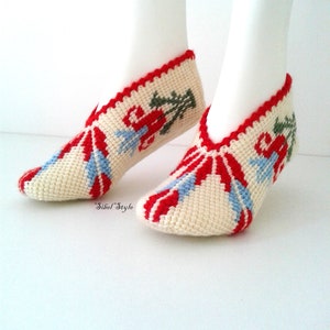 May include: A pair of cream-colored crochet slippers featuring a red, light blue, and green floral pattern. The slippers have a red scalloped trim along the opening and the text Sibel Style is printed in black near the heel area of the footwear.