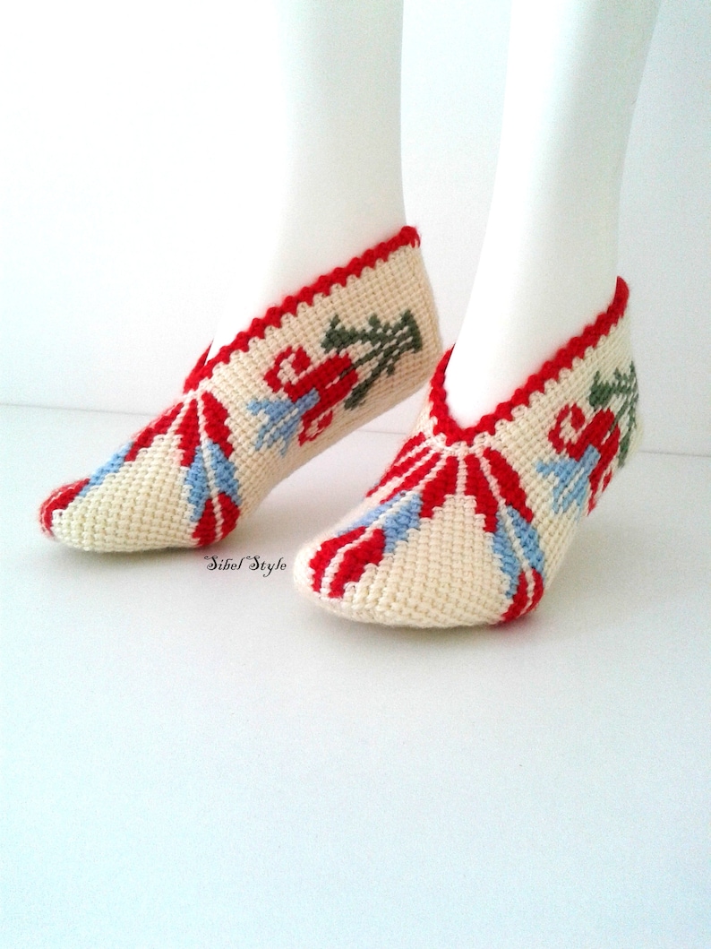 May include: A pair of cream-colored crochet slippers featuring a red, light blue, and green floral pattern. The slippers have a red scalloped trim along the opening and the text Sibel Style is printed in black near the heel area of the footwear.