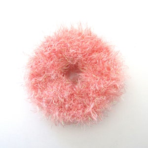 Puede incluir: Un coletero circular hecho de hilo rosa claro y difuso con fibras iridiscentes integradas que captan la luz. La textura es muy voluminosa y peluda, creando una apariencia suave y redondeada con una abertura central hueca.