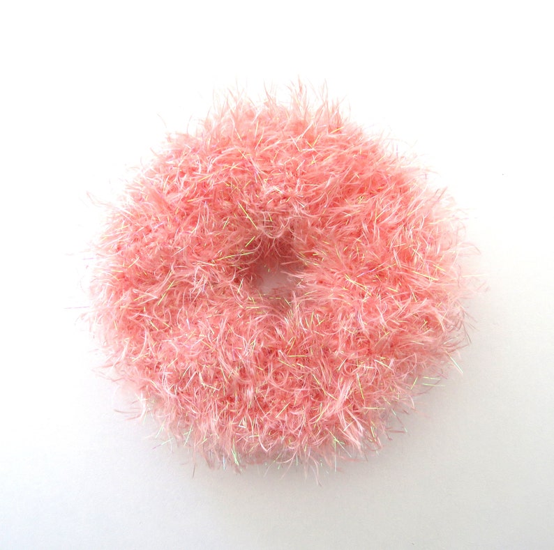 Puede incluir: Un coletero circular hecho de hilo rosa claro y difuso con fibras iridiscentes integradas que captan la luz. La textura es muy voluminosa y peluda, creando una apariencia suave y redondeada con una abertura central hueca.