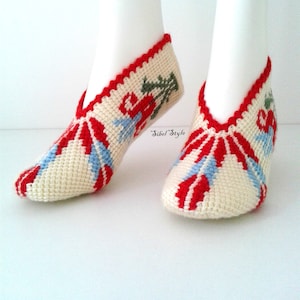 May include: A pair of cream-colored crochet slippers featuring a red and light blue sunburst pattern on the toe and a floral motif on the side. The slippers have a red crocheted trim along the opening and the text Sibel Style is visible.