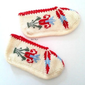 May include: A pair of off-white crochet slippers featuring a red, green, and light blue floral cross-stitch pattern. The slippers have a red crocheted trim along the top edge and the text Sibel Style printed in small black letters between them.
