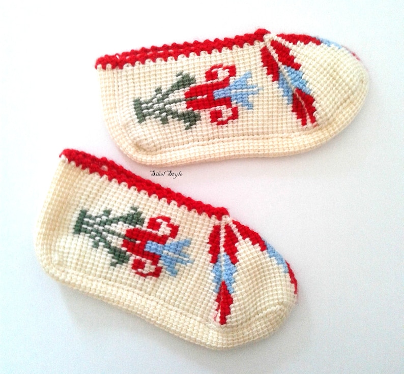 May include: A pair of off-white crochet slippers featuring a red, green, and light blue floral cross-stitch pattern. The slippers have a red crocheted trim along the top edge and the text Sibel Style printed in small black letters between them.