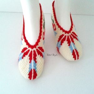 May include: A pair of off-white crochet slippers featuring a vibrant red and light blue geometric pattern. The slippers have a scalloped red trim around the opening and the text Sibel Style is printed in black script between the two slippers.