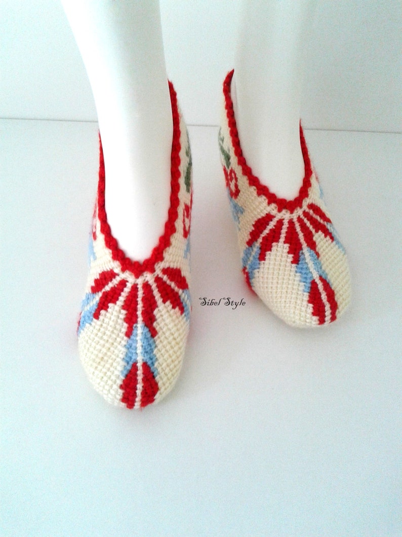 May include: A pair of off-white crochet slippers featuring a vibrant red and light blue geometric pattern. The slippers have a scalloped red trim around the opening and the text Sibel Style is printed in black script between the two slippers.