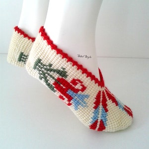 May include: A pair of cream-colored crochet slippers featuring a red, green, and light blue floral pattern. The slippers have a red crocheted trim along the opening and the text Sibel Style is printed in black near the ankle area of the footwear.