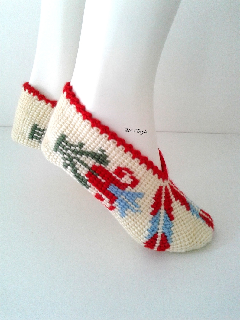 May include: A pair of cream-colored crochet slippers featuring a red, green, and light blue floral pattern. The slippers have a red crocheted trim along the opening and the text Sibel Style is printed in black near the ankle area of the footwear.