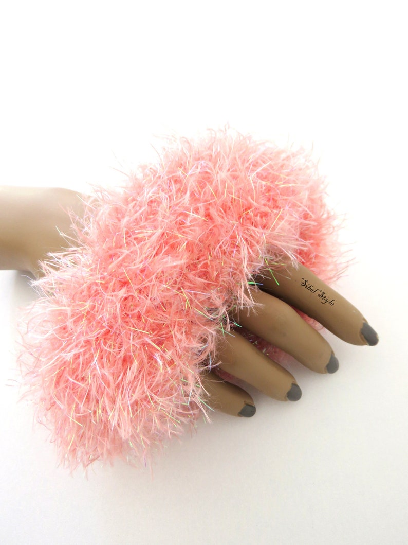 Puede incluir: Un pu&ntilde;o de mu&ntilde;eca de color rosa claro, difuso y hecho de hilo texturizado con fibras iridiscentes tejidas. El accesorio presenta una apariencia densa y peluda, exhibido en una mano de maniqu&iacute; con el texto Sibel Style en el dedo &iacute;ndice.