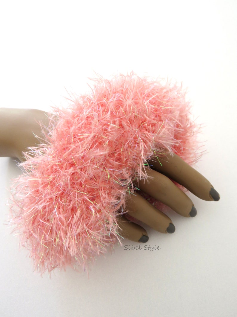 Puede incluir: Un brazalete de lana esponjosa en color rosa coral, elaborado con hilo texturizado y fibras iridiscentes entrelazadas. El accesorio presenta una apariencia densa y peluda con un acabado suave que refleja la luz. El texto Sibel Style est&aacute; impreso en negro.