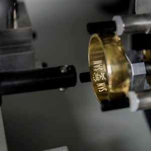 Peut inclure: Gros plan d'une bague en or en cours de gravure avec les mots "SAN X SAN" et un symbole de diamant.