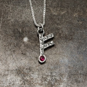 May include: Silver chain necklace with a silver pendant in the shape of the rune Fehu. The pendant has a small red gemstone in the centre.