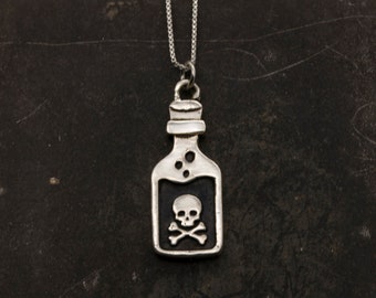 May include: A silver-toned pendant necklace featuring a bottle-shaped charm. The charm has a skull and crossbones design inside a rectangular frame. The necklace hangs on a delicate chain, perfect for adding a touch of gothic flair.