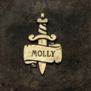 May include: Gold-toned metal pendant featuring a stylized dagger with a scroll banner that reads "MOLLY".