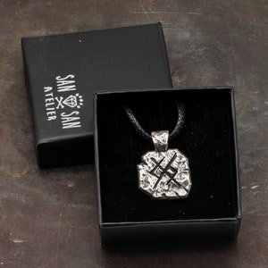 May include: A silver pendant necklace with a textured, geometric design. The pendant is in a black box with a black cord and the words "SAN SAN ATELIER" printed on the box.