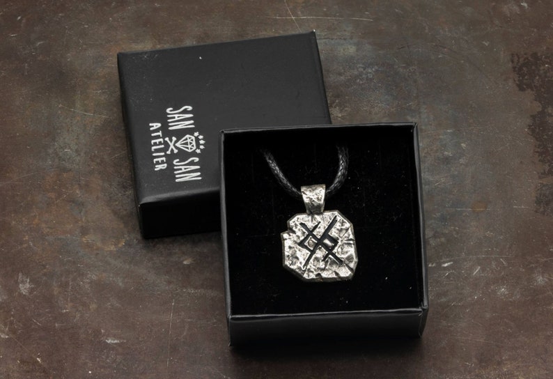 May include: A silver pendant necklace with a textured, geometric design. The pendant is in a black box with a black cord and the words "SAN SAN ATELIER" printed on the box.