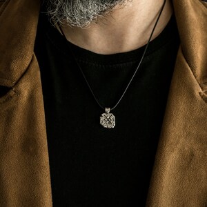 May include: A silver pendant necklace with a detailed design hangs from a black cord. The necklace is worn by a person wearing a brown suede jacket and a black t-shirt.