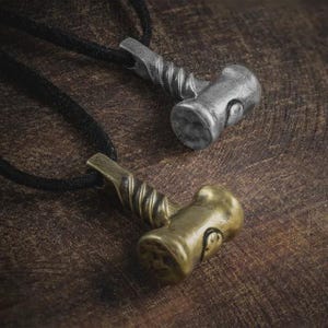 May include: Two metal Thor's hammer pendants on black cords. One is silver-toned, the other is gold-toned. The hammer heads have a textured appearance, and the handles are twisted. The pendants rest on a dark wood surface.