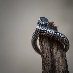 Pode incluir: Um anel de prata com um design intrincado e texturizado. O anel é enrolado em um pedaço de madeira desgastada, mostrando seus padrões detalhados e artesanato. O design do anel apresenta elementos geométricos e lineares.