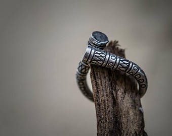 May include: A silver ring with an intricate, textured design. The ring is wrapped around a piece of weathered wood, showcasing its detailed patterns and craftsmanship. The ring's design features geometric and linear elements.